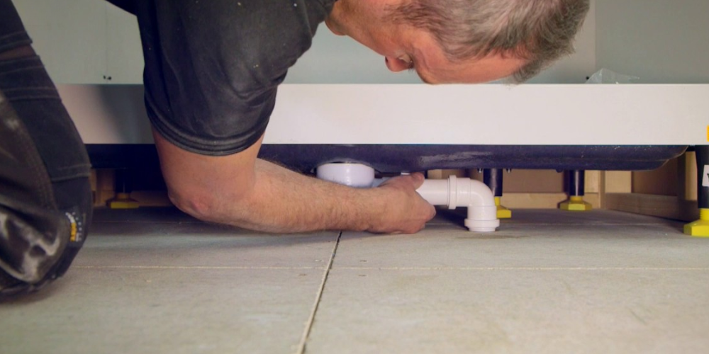 Person kneeling on tiled floor inspecting under a raised shower tray, showing precise plumbing work