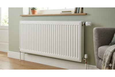 White single panel radiator mounted on a light green wall beneath a window, beside a grey sofa with a striped blanket.